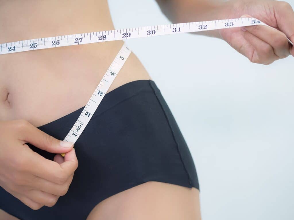 Une femme mesurant son tour de taille avec un ruban métreur noir et blanc, symbolisant l'efficacité d'un soin minceur et le remodelage de la silhouette.